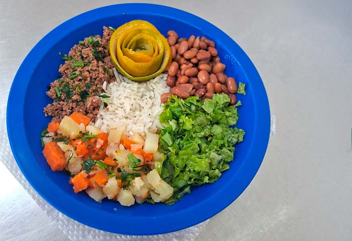 Prato saudável servido em restaurante escolar da Risotolândia, com arroz, feijão, proteína magra, legumes e fruta como sobremesa